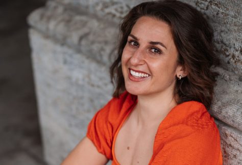 Porträt der Autorin Julie Marinello, sie sitzt an einer mauer gelehnt mit Jeans und orangenem T-Shirt